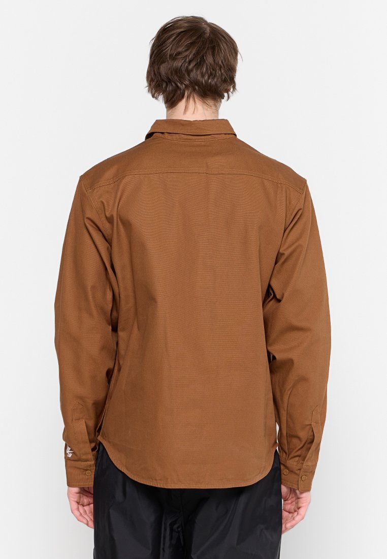 Chemise marron à manches longues avec un col boutonné et un détail de découpe au dos. Elle présente une texture lisse et des poignets avec fermetures à bouton.