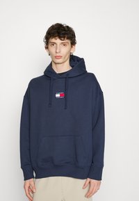 Felpa con cappuccio di colore blu navy realizzata in cotone, con tasca frontale e patch con logo che presenta accenti rossi, bianchi e blu.