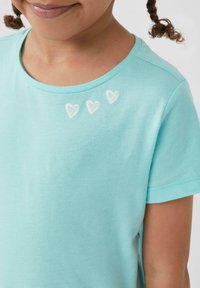 Turquoise katoenen t-shirt met een ronde hals, voorzien van drie geborduurde witte harten bij de schouder. Korte mouwen, zachte textuur.