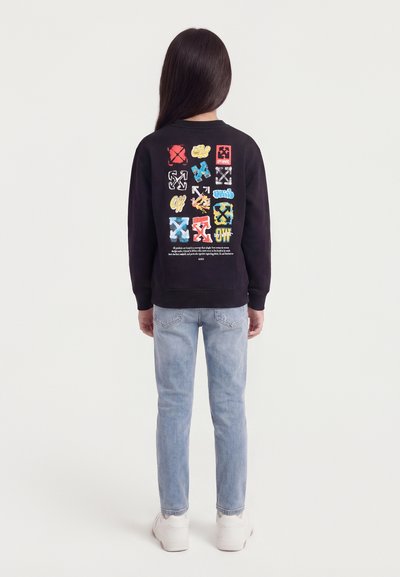 Παιδί με πλάτη που φοράει μαύρο μπλουζάκι με πολύχρωμα τετράγωνα γραφικά στο πίσω μέρος, ανοιχτόχρωμο μπλε τζιν και λευκά αθλητικά παπούτσια.