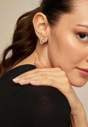 Pendientes de plata con una forma ovalada abstracta que presenta un acento interior rosa. Llevados por una modelo de cabello oscuro y blusa negra, destacando la textura y el diseño.