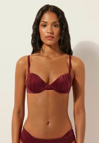 Góra od bikini w kolorze fioletowym z usztywnianymi miseczkami, plisowaną fakturą i skręconym węzłem na środku. Cienkie regulowane ramiączka łączą się z jednolitym pasem.