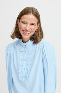 Hellbluse aus glattem Stoff, mit einem gerafften Vorderteil, Knopfverschluss und Puffärmeln. Dünner Kragen mit dezentem Glanz.