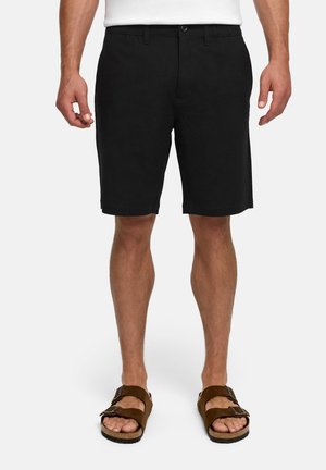 Homme portant un short noir jusqu'aux genoux et des sandales marron à boucle, debout devant un fond blanc.