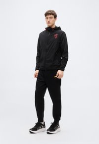 Μαύρο ελαφρύ μπουφάν με κουκούλα και κόκκινες λεπτομέρειες, συνδυασμένο με μαύρο φόρμα παντελόνι με κίτρινο λογότυπο. Μαύρα αθλητικά παπούτσια ολοκληρώνουν την εμφάνιση.