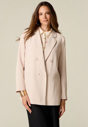 Frau mit langen braunen Haaren, die einen beigen doppelt geknöpften Blazer über einer geblümten Bluse und schwarzen Hosen trägt, steht vor einem neutralen Hintergrund.