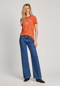 T-shirt orange à manches courtes avec un график blanc indiquant "Jeans 73", associé à un jean en denim large bleu et des bottes à talons marron.