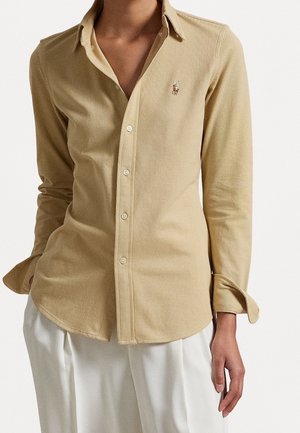 Chemise beige à manches longues boutonnée avec un petit logo de cheval brodé sur la poitrine, portée avec un pantalon plissé blanc.