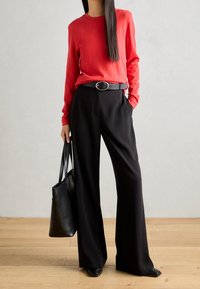 Camicia rossa a maniche lunghe, pantaloni neri a gamba larga, cintura nera con fibbia circolare, borsa nera e scarpe a punta, su pavimento di legno.