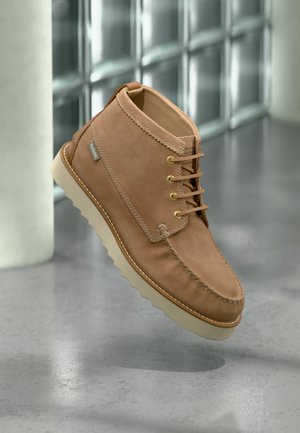 Lichtbruine suède enkellaars met beige geribbelde zool, gouden veteroogjes, leren veters en een zichtbare Barbour merklabel aan de zijkant.