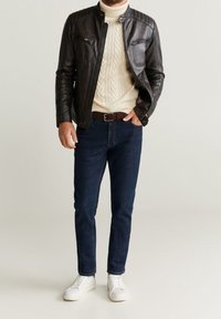 Hnědá kožená bunda s detailním prošíváním, krémový svetr s copánkovým vzorem, tmavě modré džíny a bílé tenisky.