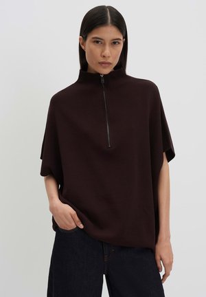 Jonge vrouw met lang donker haar draagt een losse donkerbruine top met halflange mouwen en een rits aan de voorkant, gecombineerd met een donkere broek.