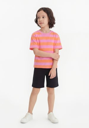 Παιδί που στέκεται με σταυρωμένα τα χέρια, φοράει ριγέ ροζ και πορτοκαλί μπλούζα, μαύρο σορτς και λευκά αθλητικά παπούτσια σε λευκό φόντο.