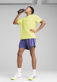 Gelbes Sport-T-Shirt und lila Shorts, kombiniert mit weißen und neonfarbenen Schuhen, h�ält eine schwarze Wasserflasche. Schlichte, funktionale Sportbekleidung.