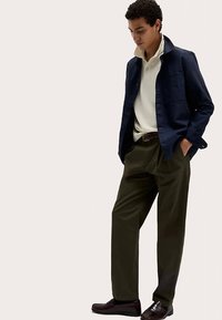 Marinblå bomullsjacka med knappar över en krämfärgad pikétröja, kombinerad med olivgröna plisserade byxor och mörkbruna läderskor.