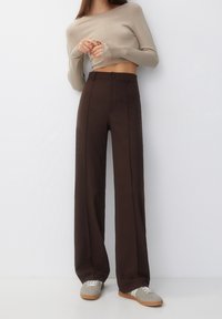 Bruine wijde pantalons met plooien aan de voorkant, knoopsluiting en een gladde afwerking, gecombineerd met een beige kortere top met lange mouwen en grijze schoenen.