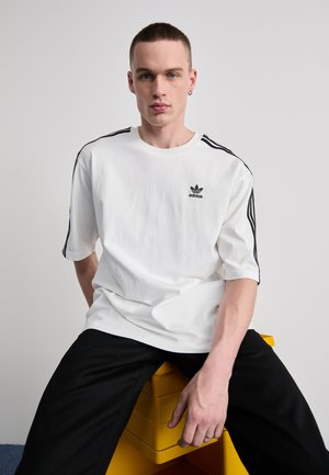 White cotton t-shirt featuring short sleeves, black three-stripe design along shoulders, and a small black Adidas logo on the chest.