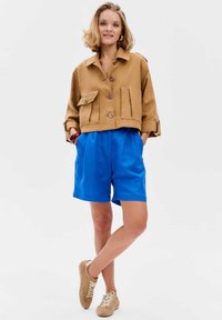Tan cropped jack met twee voorzakken en grote knopen, gecombineerd met blauwe plooishorts en beige sneakers met witte veters.