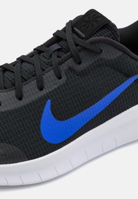 Chaussure de sport noire avec tige en tissu texturé, présentant un logo Nike swoosh bleu bien visible, des lacets noirs et une semelle blanche dentelée.