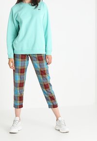 Una persona indossa un felpa teal, pantaloni cropped a quadretti in rosso, blu e giallo, e sneaker chunky bianche, in piedi con le gambe divaricate su uno sfondo bianco.