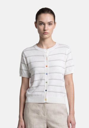 Jeune femme aux cheveux attachés, portant un cardigan blanc à manches courtes avec des rayures grises et des boutons multicolores, assorti à un pantalon beige clair.