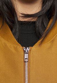 Musztardowa kurtka bomberka z prążkowanym kołnierzem i widocznym srebrnym suwakiem, noszona na czarnej, fakturowanej koszuli.