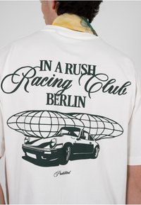 Hvit T-skjorte med et svart grafisk bilde av en bil med et jordklode-design over. Teksten lyder «IN A RUSH Racing Club BERLIN» og «Forbudt.»