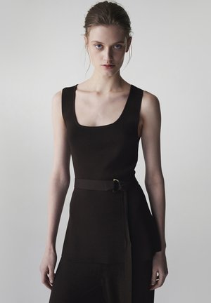 Giovane donna con i capelli raccolti, che indossa un abito nero senza maniche con scollo quadrato e una cintura abbinata, in piedi davanti a uno sfondo grigio uniforme.