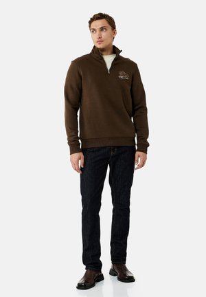 Hombre con suéter marrón oscuro con cierre parcial y diseño bordado, vaqueros negros y botas de cuero marrón oscuro, de pie frente a un fondo blanco.