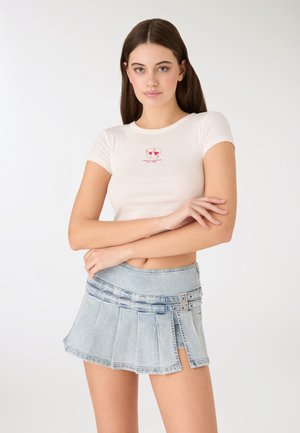 Top corto bianco a maniche corte con grafica e testo rossi, abbinato a una minigonna plissettata in denim azzurro chiaro con bottoni decorativi e accessori in metallo.