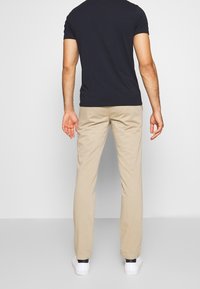 Pantaloni di cotone beige con design a gamba dritta, indossati con una camicia a maniche corte blu navy e sneakers bianche, visti da dietro.