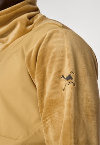 Cuello de tortuga de pana beige con una mezcla de tejidos suave, textura acanalada y un emblema de camello bordado en negro en la manga izquierda.