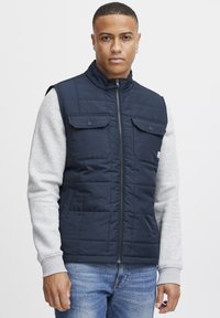 Marineblaues gesteppte ärmelloses Weste mit zwei frontalen Taschen, grauen langen Ärmeln und einem Reißverschluss. Glattes Material mit matter Oberfläche.