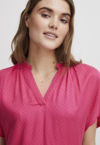 Blusa rosa con un delicato motivo a diamante, scollatura a V con dettagli a volant e maniche corte in un tessuto leggero e testurizzato.