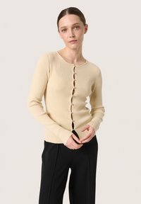 Ribbad beige kofta med långa ärmar, rund nacke och knappstängning som har dekorativa knappar. Buren över svarta byxor.