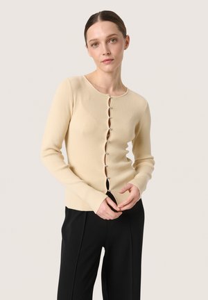 Ribbed beige cardigan with long sleeves, round neck, and button closure featuring decorative buttons. Worn over black trousers.