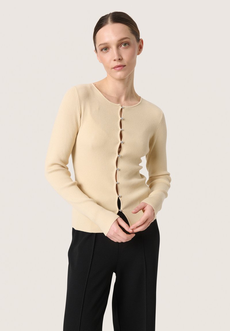 Cardigan beige a coste con maniche lunghe, collo rotondo e chiusura con bottoni decorativi. Indossato sopra pantaloni neri.