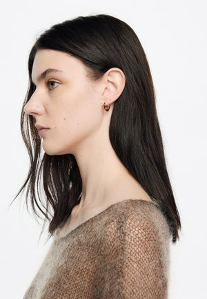 Pendiente de aro dorado en una modelo, presentado con un suéter suave, texturizado y de color marrón claro. El diseño presenta una forma simple y redondeada.