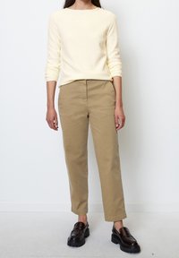 Pull texturé crème à manches longues, associé à un pantalon chino marron clair fuselé et des mocassins foncés, sur un fond uni.