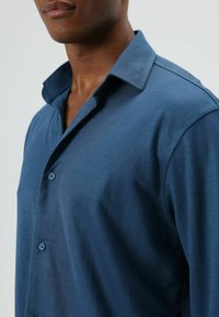 Overhemd met knopen in donkerblauwe stof. Heeft een klassieke kraag, een gladde textuur en een knoopsluiting met één rij knopen. Strakke, getailleerde pasvorm.