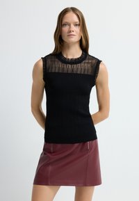 Czarny bezrękawnik z teksturowanym prążkowaniem i przezroczystą górną częścią. Połączony z burgundową skórzaną mini spódnicą z zamkami błyskawicznymi.