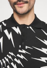 Homme portant une chemise noire à col avec un motif d'éclair blanc et trois boutons noirs, montrant le bas du visage et le cou avec une barbe courte.