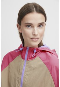 Chaqueta a bloque de color que presenta tonos rosa, beige y lavanda, fabricada en material ligero con cierre de cremallera frontal y capucha ajustable.
