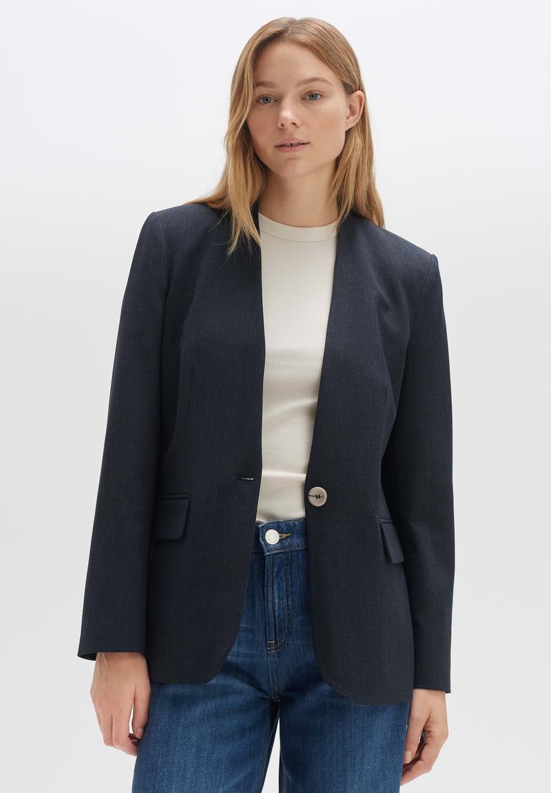 Blazer bleu marine foncé en tissu lisse, avec une fermeture à un bouton, des revers crantés et deux poches avant. Associé à un haut clair.