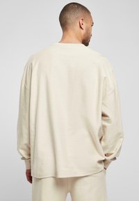 Beige geribbelde long sleeve top met een relaxte pasvorm, verlaagde schoudernaden en een zachte textuur, weergegeven vanaf de achterkant met bijpassende broek.