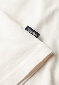 Camiseta de algodón blanca con una etiqueta de tela azul marino que lleva la palabra "Superdry". Primer plano de la manga y los detalles de la costura.