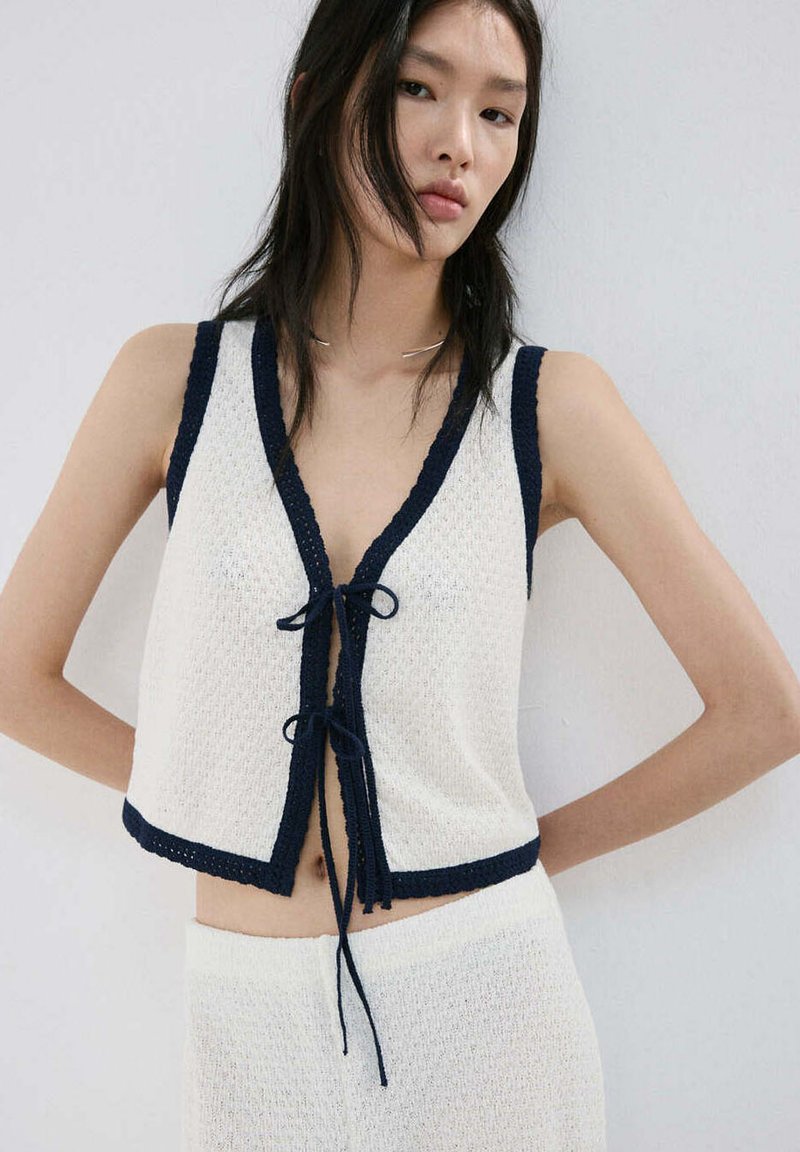 Femme aux longs cheveux foncés portant un top blanc sans manches en maille avec bordure noire et jupe assortie, debout contre un mur clair uni.