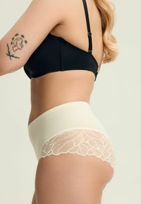 Beżowe majtki z wysokim stanem, ozdobione skomplikowaną koronką przy otworach na nogi, w połączeniu z czarnym biustonoszem. Gładki materiał, elegancki design.