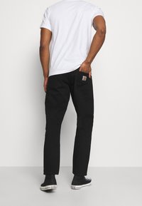 Pantaloni in denim nero con taglio dritto, dotati di una tasca posteriore e una patch con logo. Indossati con una t-shirt bianca semplice e sneakers nere.