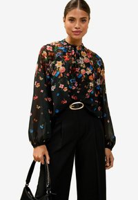 Blusa nera con fantasia floreale caratterizzata da colorati motivi di fiori, scollatura arricciata e maniche lunghe a palloncino. Abbinata a pantaloni neri a vita alta e una borsa.